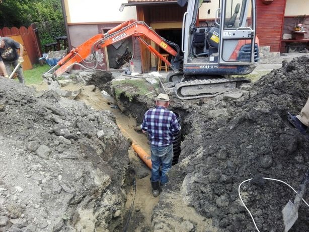 Wykop pod rury kanalizacyjne z pomarańczową rurą w tle, koparka Kubota KX41-2 VC i dwóch robotników przy pracy.