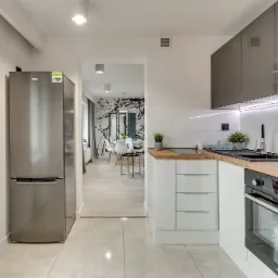 Jasna kuchnia z szarymi szafkami wiszącymi, drewnianym blatem, białymi szafkami dolnymi i srebrną lodówką, widok na jadalnię z abstrakcyjną tapetą