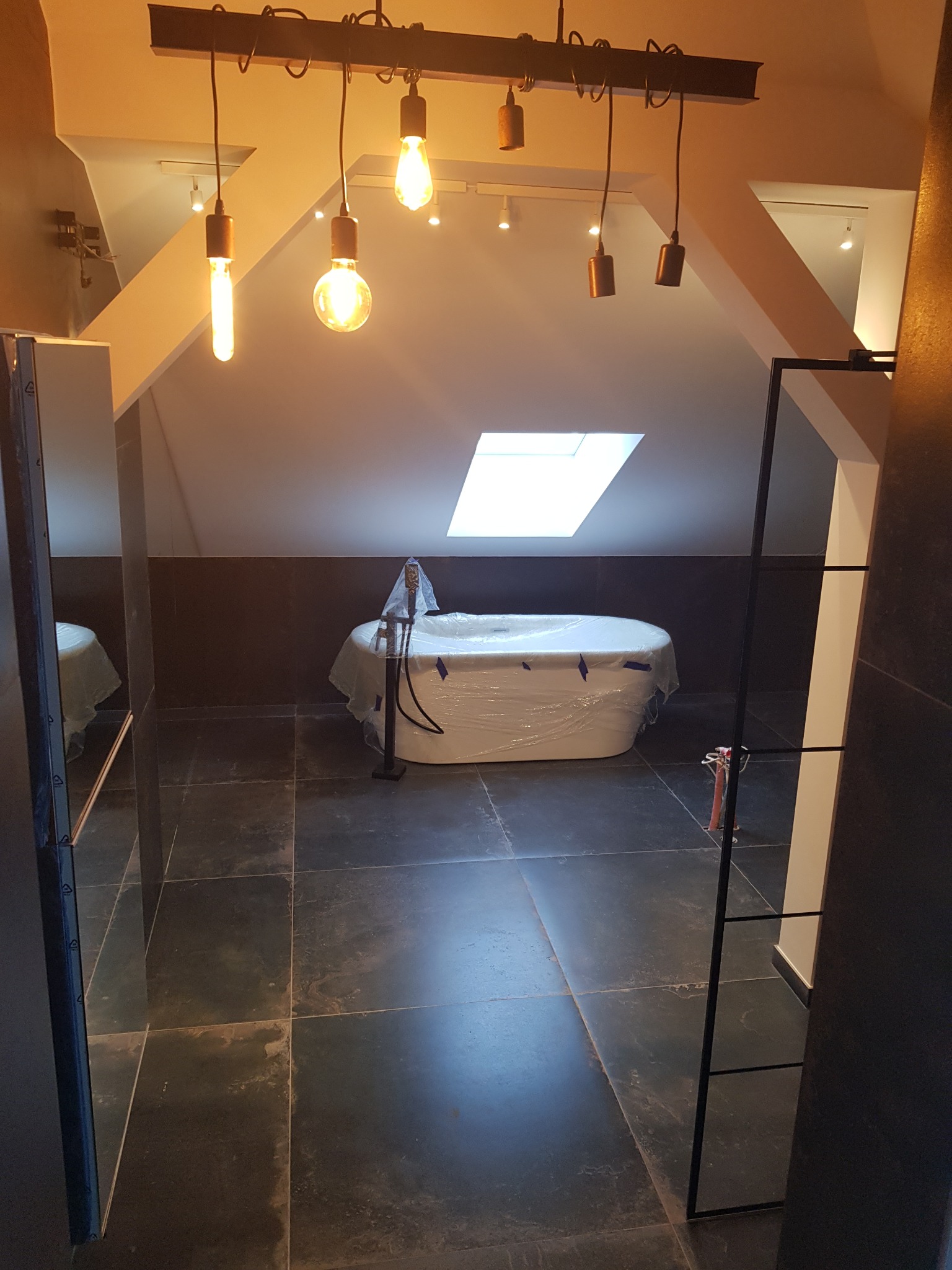 Nowoczesna łazienka na poddaszu z wanną owiniętą folią ochronną, czarnymi płytkami na ścianach i podłodze oraz wiszącymi lampami w stylu industrialnym.