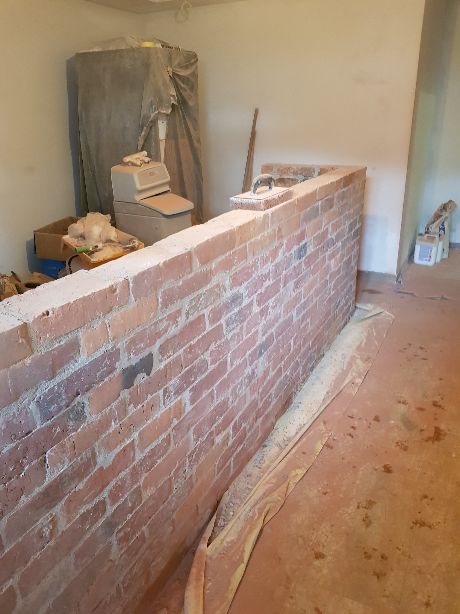 Wznoszenie niskiej ścianki z cegły w surowym wnętrzu, widoczne narzędzia murarskie na szczycie, zabezpieczona podłoga folią malarską.