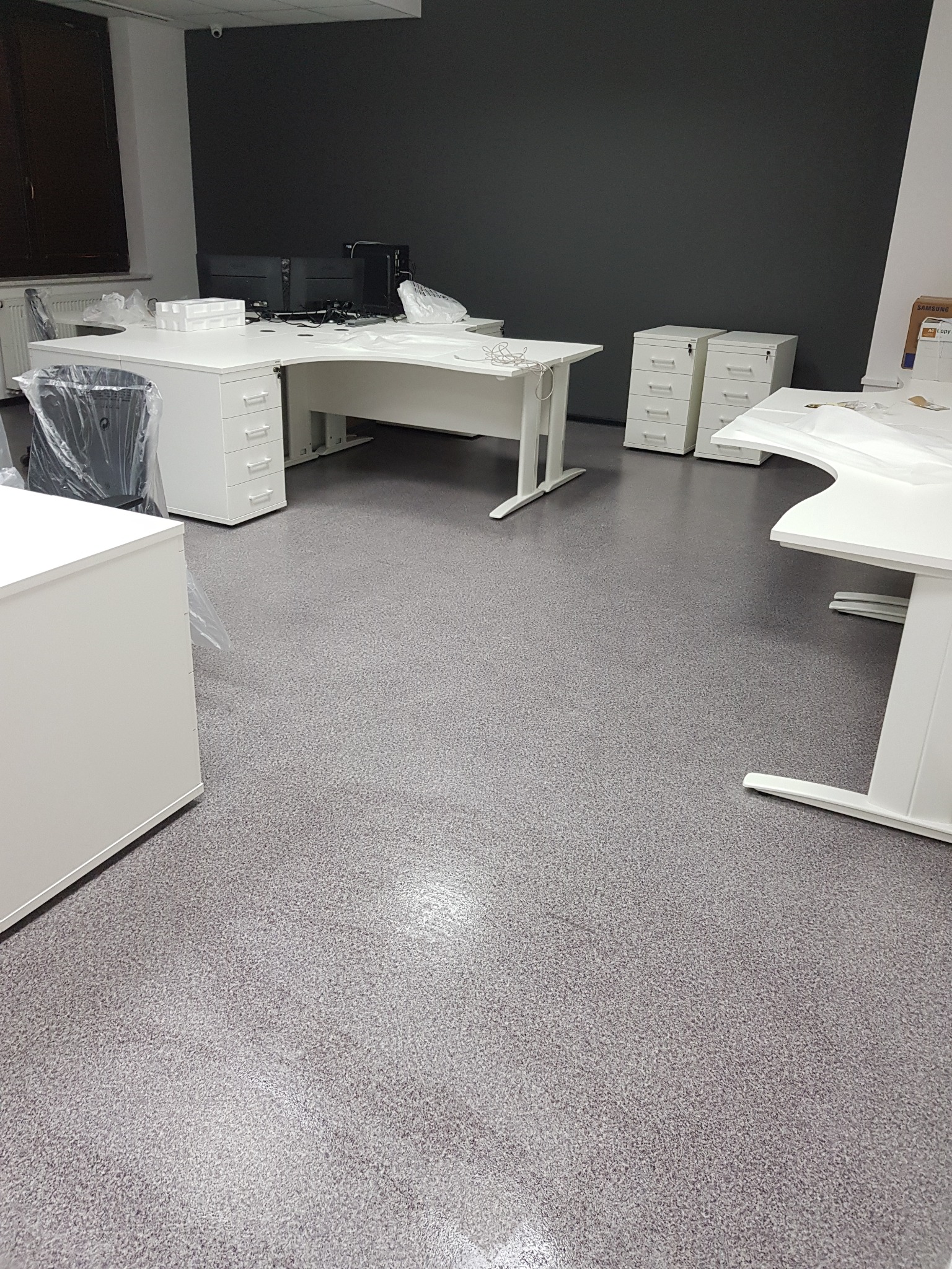 Nowoczesne biuro z białymi biurkami i szafkami, krzesła w folii ochronnej, szara podłoga i ściana akcentująca, świeżo po remoncie lub montażu.