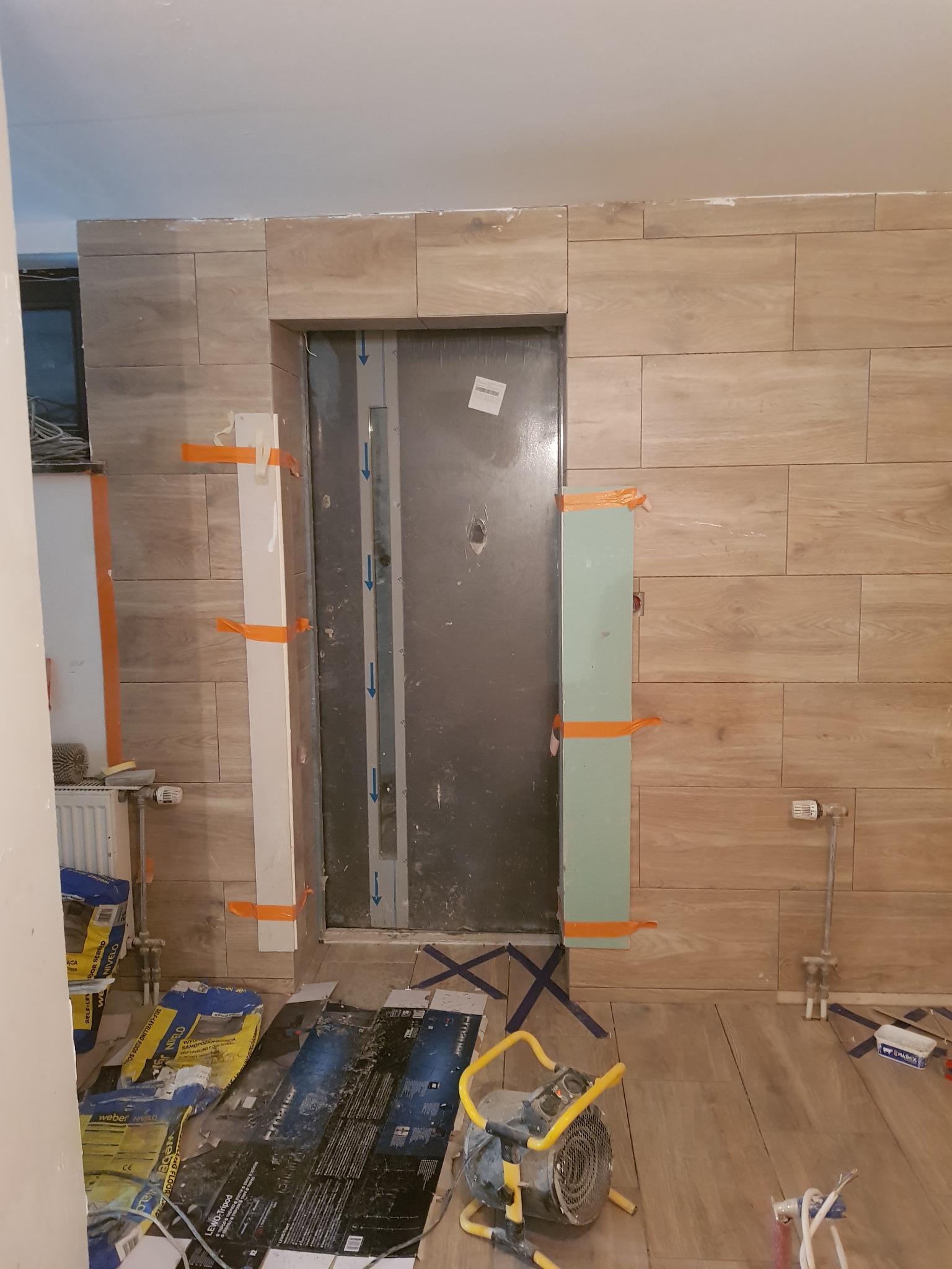 Montaż płytek imitujących drewno na ścianie wokół futryny drzwiowej, z widocznymi materiałami budowlanymi i narzędziami pracy na podłodze.