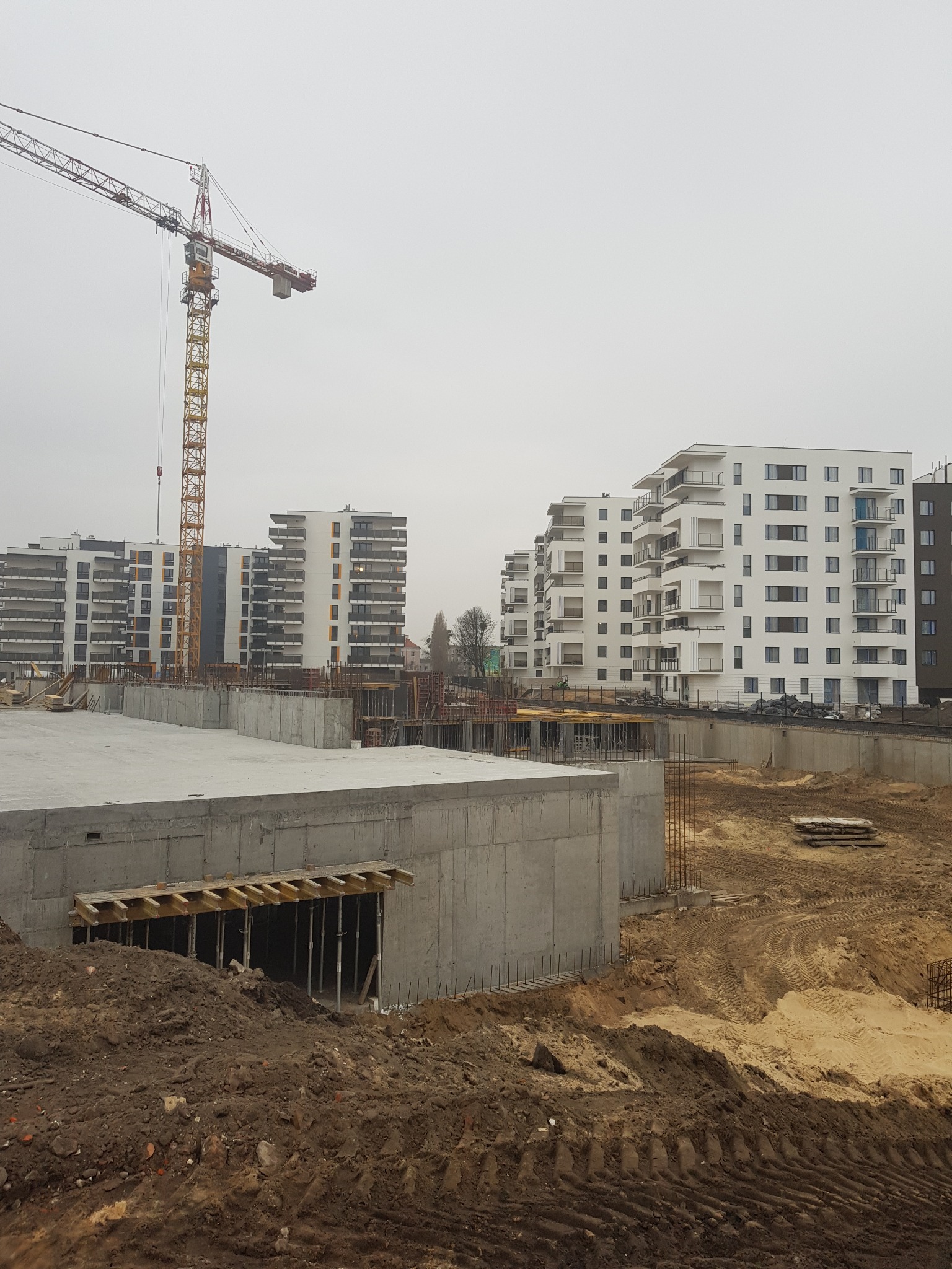 Budowa osiedla mieszkaniowego z widocznym żurawiem budowlanym, surowym betonem, i przygotowanym terenem pod dalsze prace.
