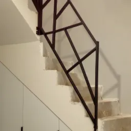 Metalowa, geometryczna balustrada schodowa w ciemnym kolorze, zamontowana na surowych, betonowych stopniach. Pod schodami zabudowa szafy wnękowej z białymi frontami i pionowymi, minimalistycznymi...