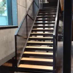 Metalowe schody z drewnianymi stopniami w nowoczesnym wnętrzu, widoczne z dołu, z czarną ramą i siatką zabezpieczającą, obok betonowa ściana z oknem.