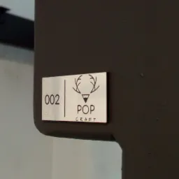 Metalowa tabliczka z numerem '002' i logo 'POP CRAFT' przymocowana do ciemnego, matowego elementu mebla.