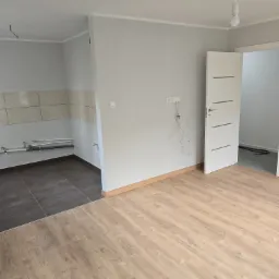 Wnętrze w trakcie wykańczania: widoczne panele podłogowe, szare ściany z białą listwą, białe drzwi wewnętrzne, fragment kuchni z ciemnymi płytkami i instalacją hydrauliczną oraz wystające kable...