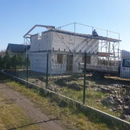 Budowa domu jednorodzinnego z białych bloczk&oacute;w, widoczne rusztowanie, drewniana konstrukcja dachu i pracownik na g&oacute;rze, siatka ogrodzeniowa w pierwszym planie.