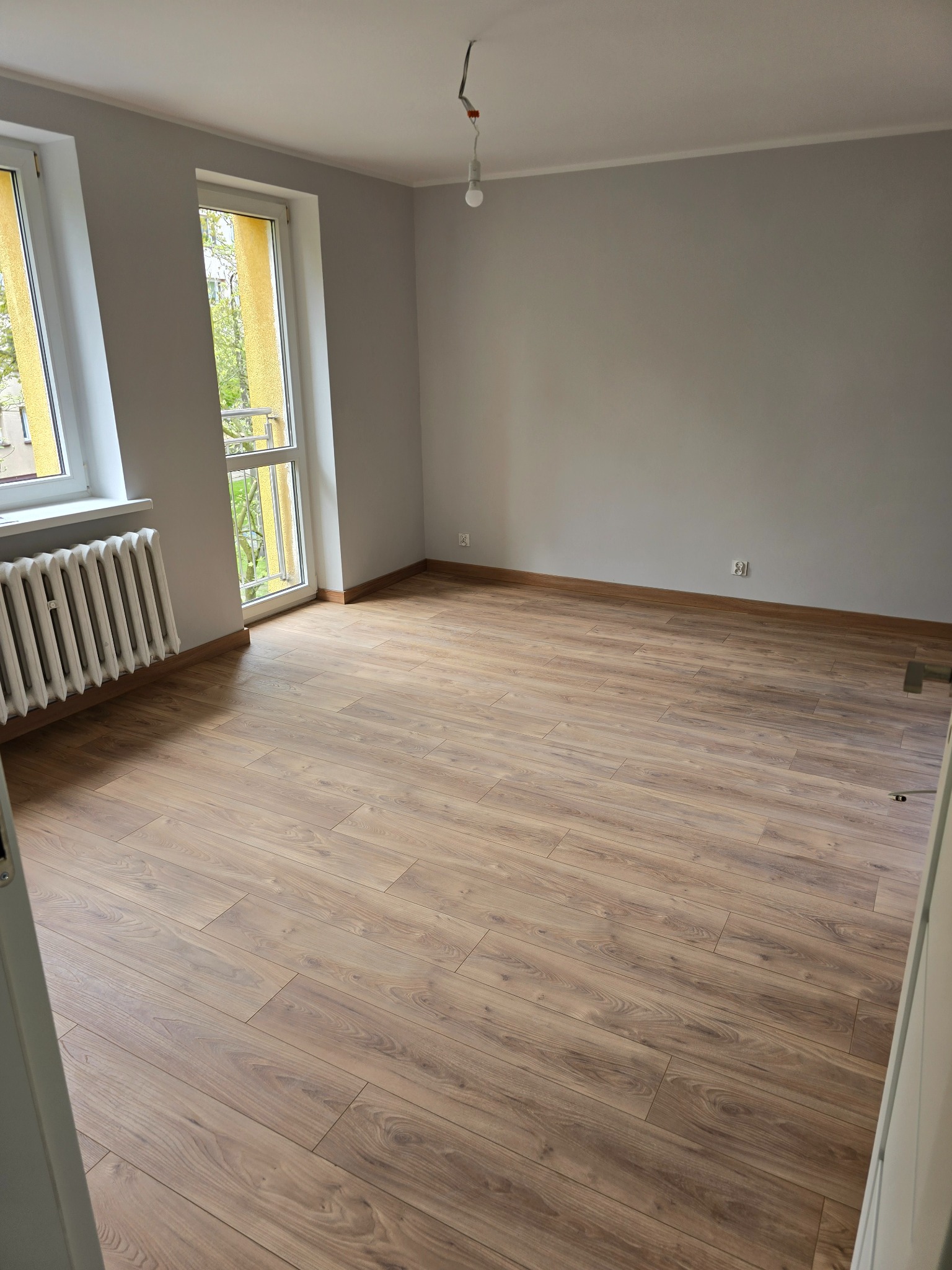 Pusty pokój po remoncie z nową podłogą z paneli imitujących drewno, szarymi ścianami, białymi oknami i grzejnikiem żeliwnym.