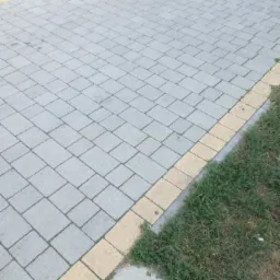 Ułożona szara kostka brukowa z obrzeżem w kolorze piaskowym, przylegająca do trawnika z widocznymi źdźbłami trawy.