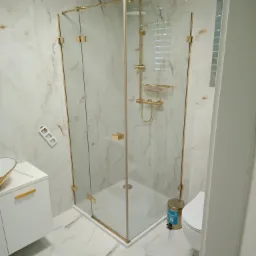 Nowoczesna kabina prysznicowa z złotymi elementami wykończeniowymi, marmurowe płytki na ścianach i podłodze, biała szafka umywalkowa z złotymi uchwytami, widoczny kosz na śmieci i toaleta.