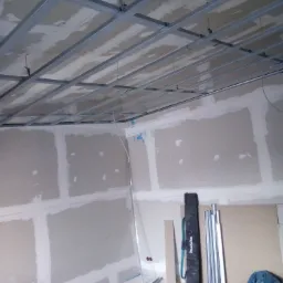 Montaż metalowego stelażu sufitowego i płyt gipsowo-kartonowych na ścianach w trakcie prac wykończeniowych, widoczne narzędzia i materiały budowlane.