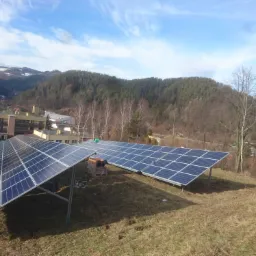 Dwa rzędy paneli słonecznych na metalowych konstrukcjach, na tle pagórkowatego krajobrazu z budynkami i lasem, w słoneczny dzień.