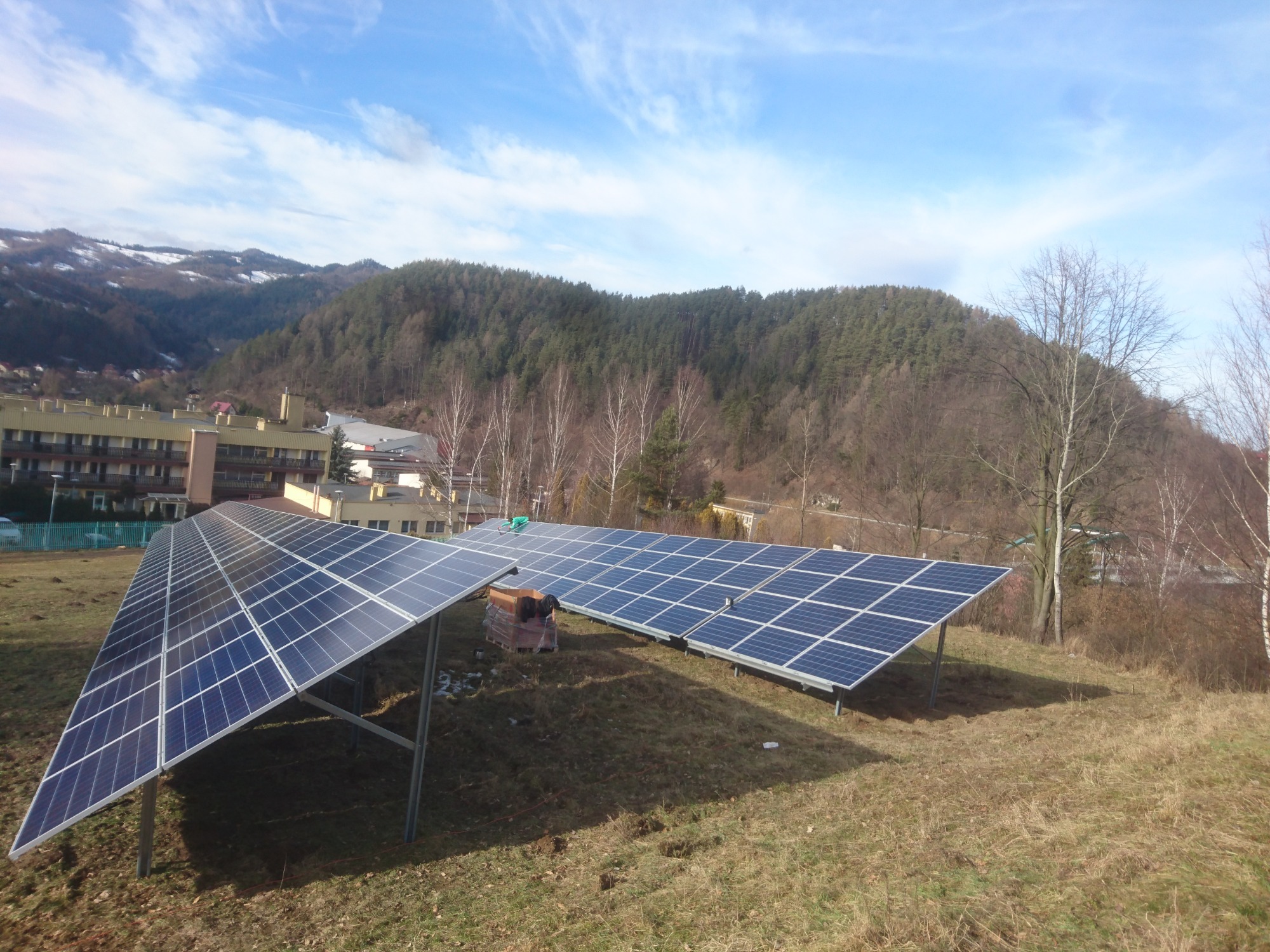 Dwa rzędy paneli słonecznych na metalowych konstrukcjach, na tle pagórkowatego krajobrazu z budynkami i lasem, w słoneczny dzień.