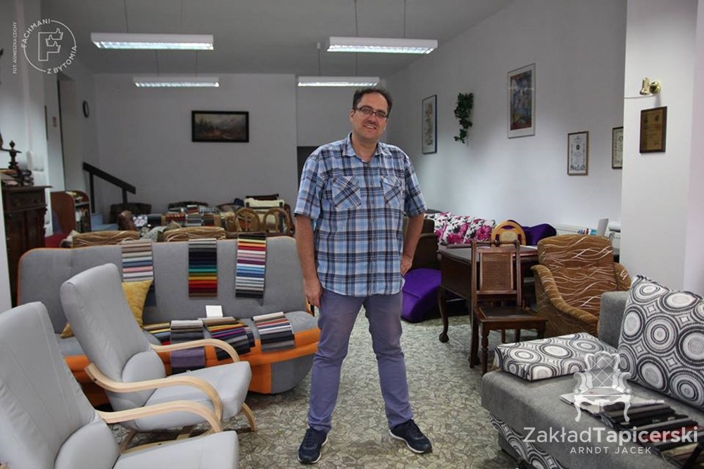 Wnętrze zakładu tapicerskiego z pracownikiem na pierwszym planie, prezentacja próbek tkanin na meblach, widoczne fotele, sofy i krzesła w różnych stylach.