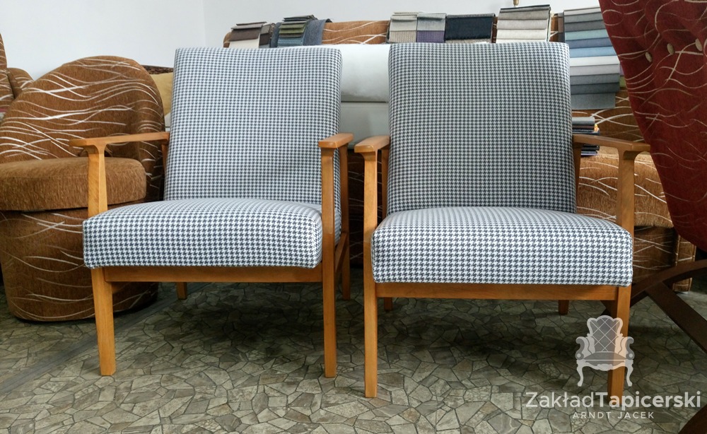 Dwa identyczne fotele tapicerowane tkaniną w pepitkę, drewniane nóżki i podłokietniki, w tle próbki tkanin i inne meble, widoczne logo zakładu tapicerskiego.