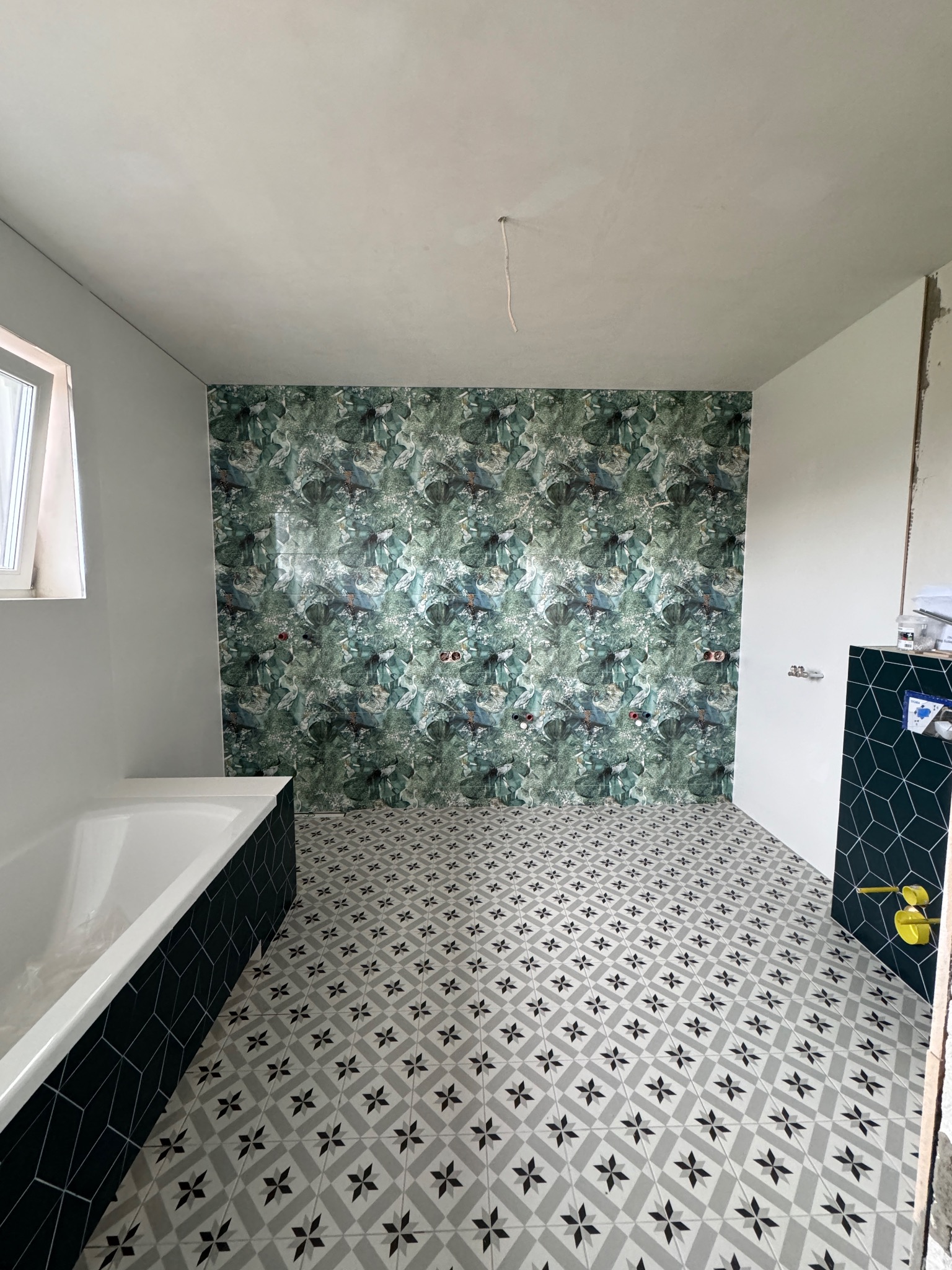 Łazienka w trakcie remontu: wzorzysta tapeta z motywem roślinnym na ścianie, płytki podłogowe w geometryczny wzór, obudowa wanny z ciemnych płytek, widoczne niezabezpieczone kable elektryczne...