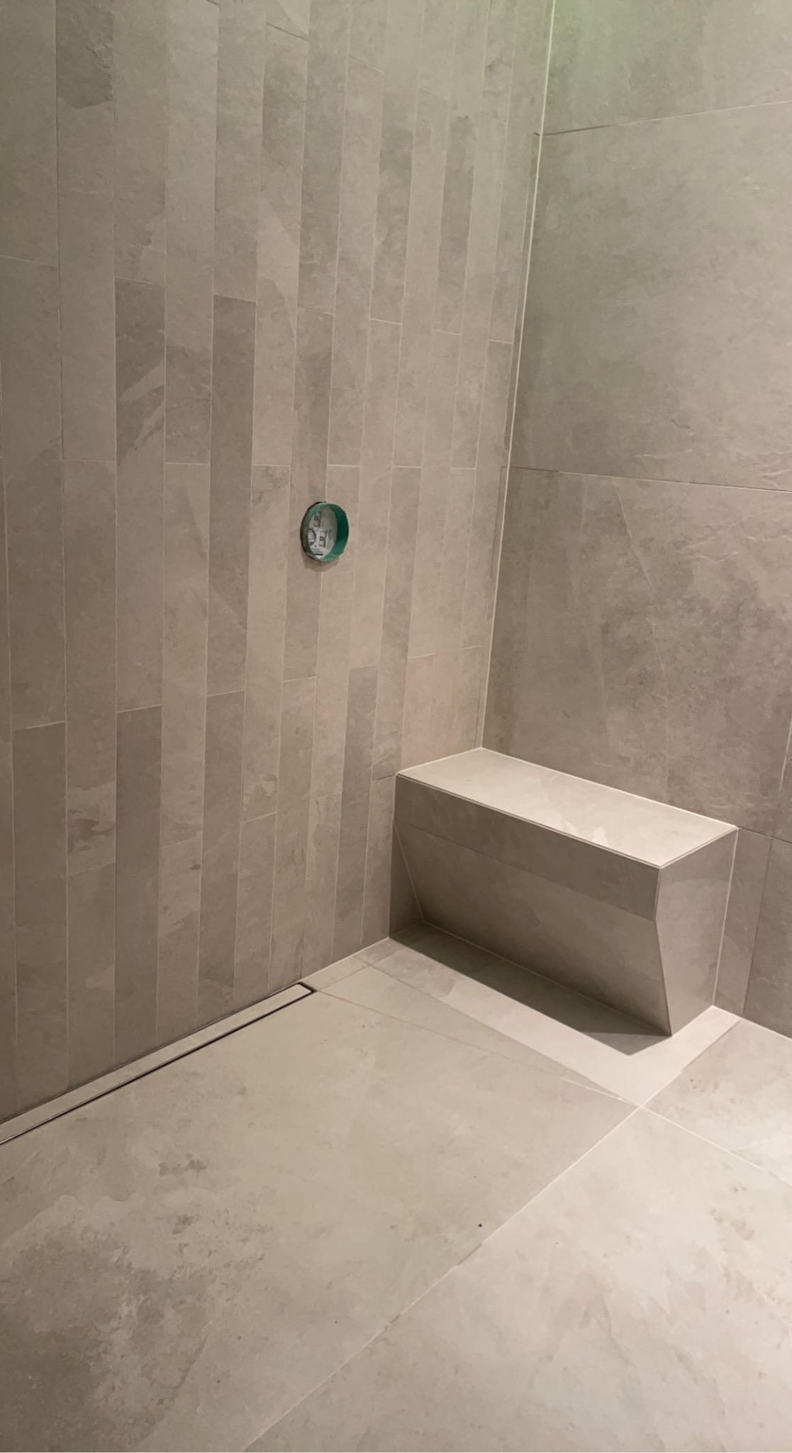 Fragment łazienki z beżowymi, podłużnymi płytkami na ścianie, siedziskiem z płytek i odpływem liniowym w podłodze, widoczny otwór na baterię.