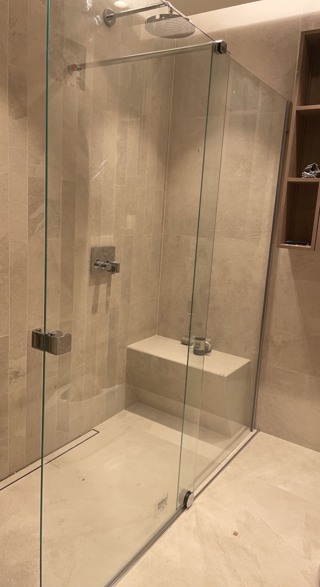 Nowoczesna kabina prysznicowa typu walk-in z siedziskiem, wykończona beżowymi płytkami imitującymi kamień, z widoczną armaturą i odpływem liniowym w podłodze. Na półce widoczny kubek i pojemnik.