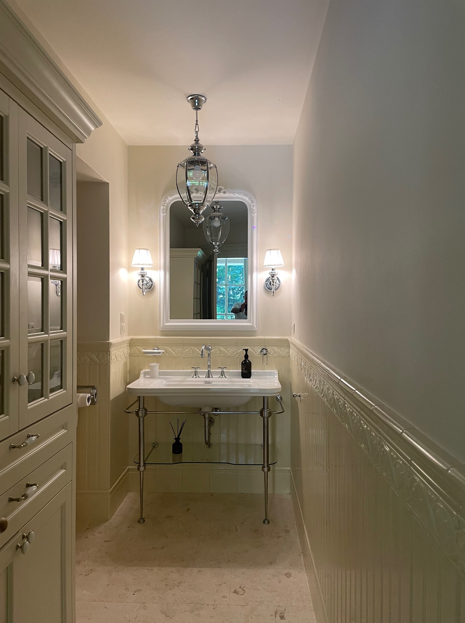 Jasna łazienka w stylu klasycznym z umywalką na metalowych nogach, ozdobnymi płytkami ściennymi, lustrem w białej ramie i chromowanymi kinkietami.