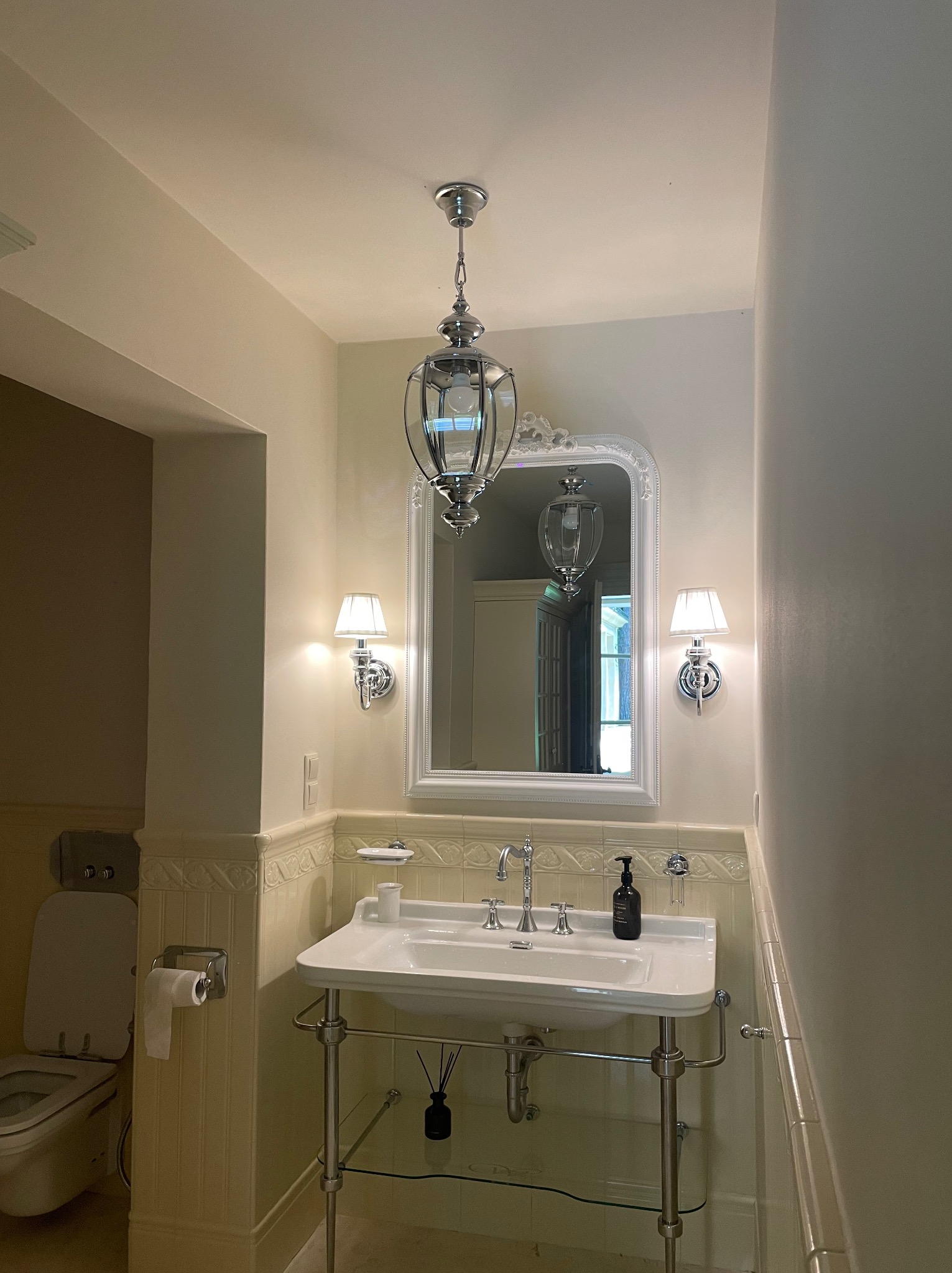 Elegancka łazienka z umywalką na metalowych nogach, ozdobnym lustrem i chromowanymi kinkietami, oświetlona wiszącą lampą w stylu retro.