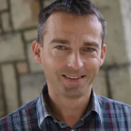 Portret uśmiechniętego mężczyzny w koszuli w kratę na tle ściany z kamienia.