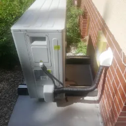 Zewnętrzna jednostka pompy ciepła Kaisai zamontowana przy ceglanej ścianie budynku, widoczne przyłącza i otoczenie z zielenią.