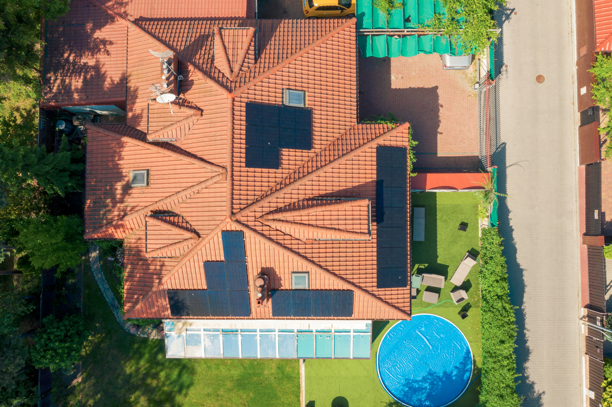 Widok z drona na dom z czerwoną dachówką i panelami słonecznymi, basen z boku i zielony trawnik.