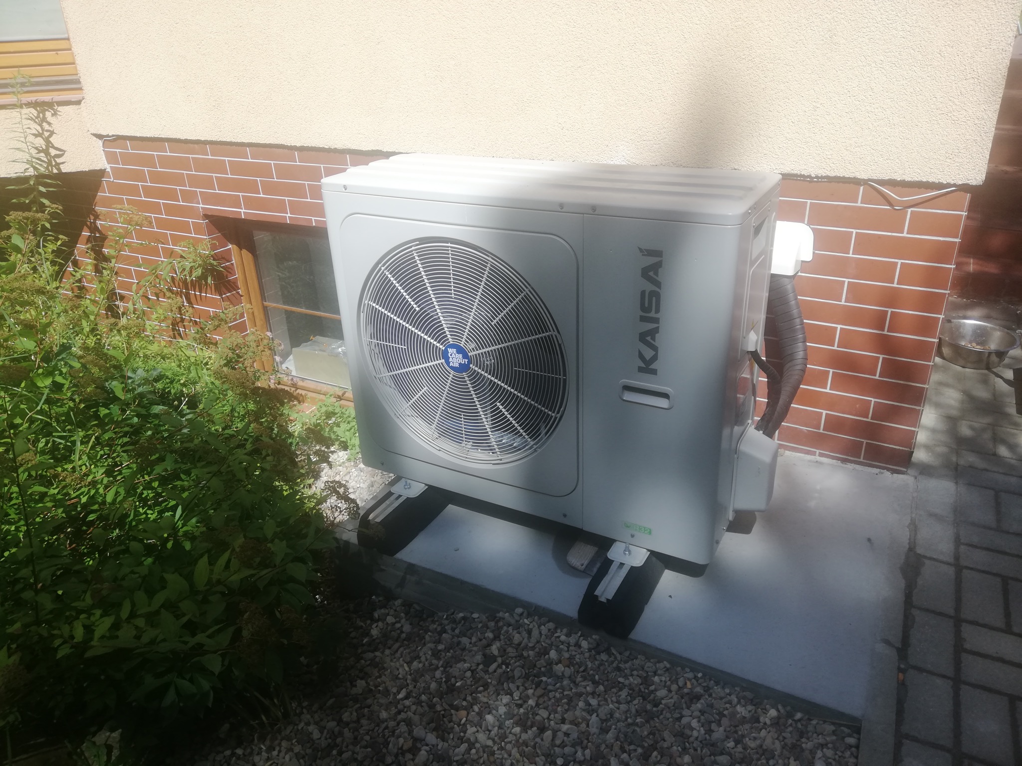 Zewnętrzna jednostka pompy ciepła marki Kaisai, zamontowana na betonowej podstawie przy ścianie budynku z ceglaną elewacją, widoczna zieleń i kamyki.