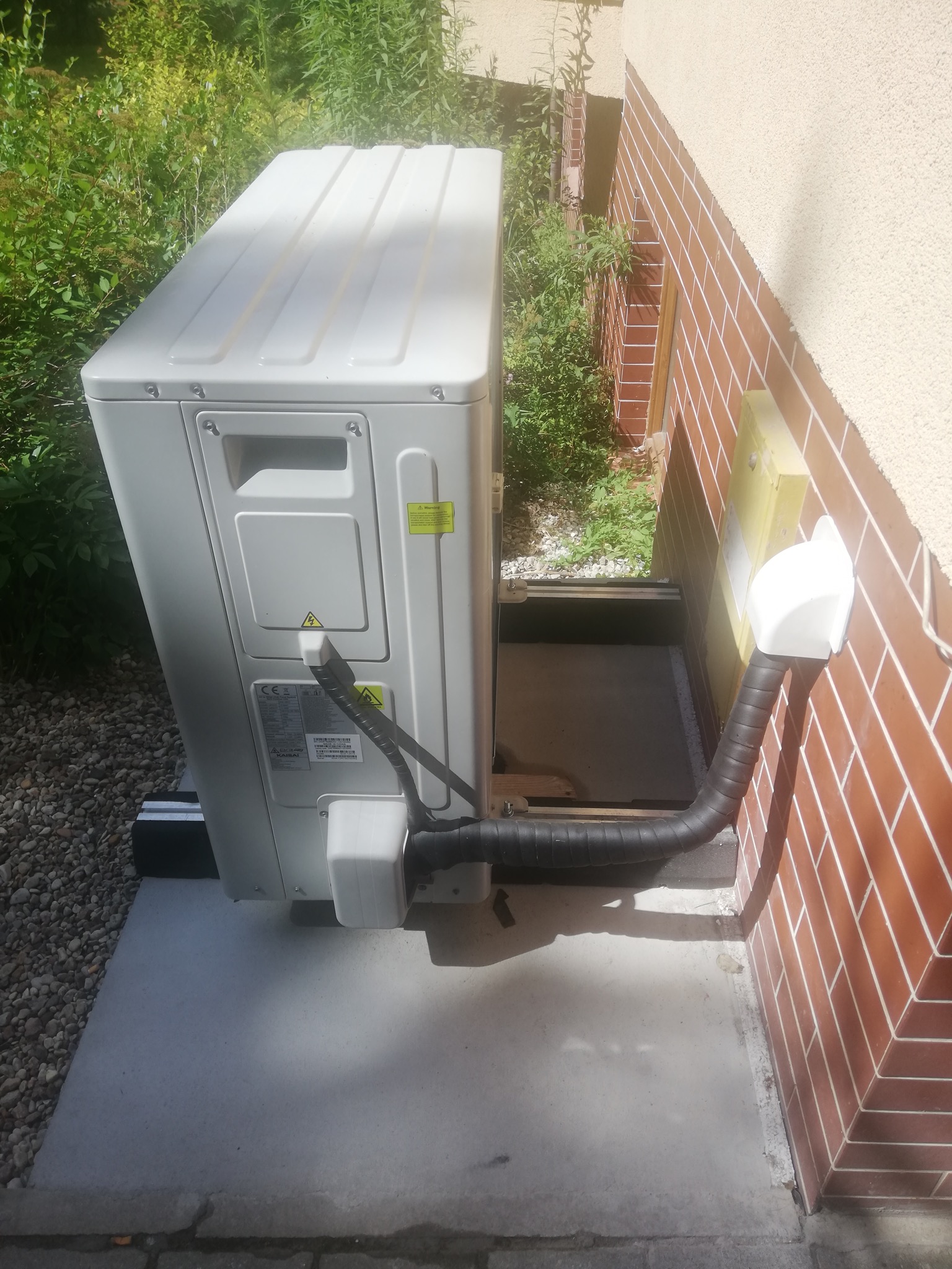 Zewnętrzna jednostka pompy ciepła Kaisai zamontowana przy ceglanej ścianie budynku, widoczne przyłącza i otoczenie z zielenią.