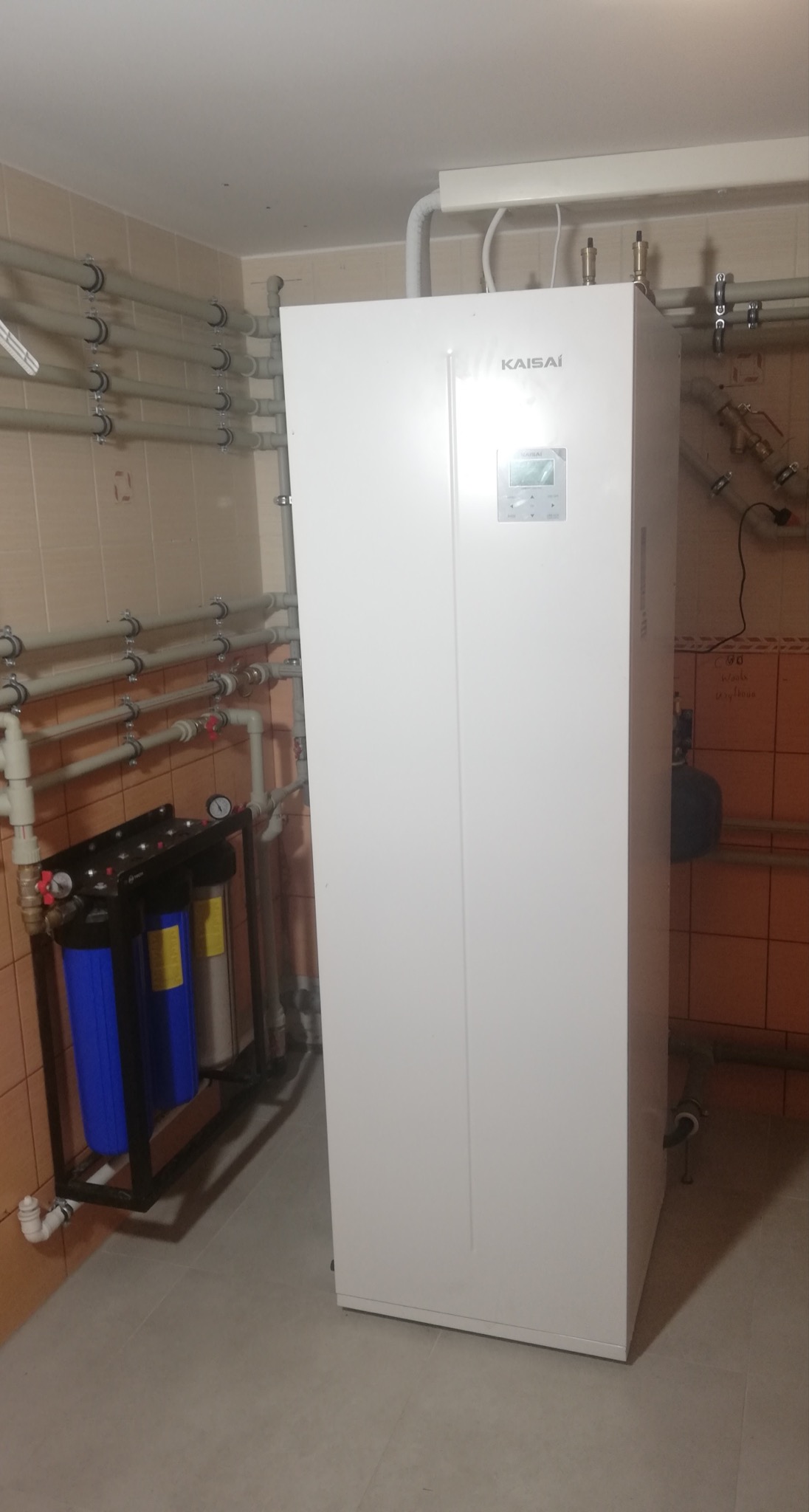 Biała pompa ciepła marki Kaisai zainstalowana w pomieszczeniu z beżowymi płytkami, widoczne rury i filtry wody.
