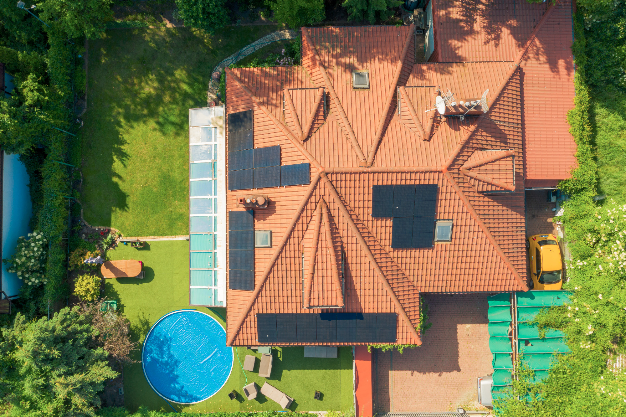 Widok z lotu ptaka na dom z panelami słonecznymi na dachu, basenem z niebieską wodą i żółtym samochodem zaparkowanym obok.