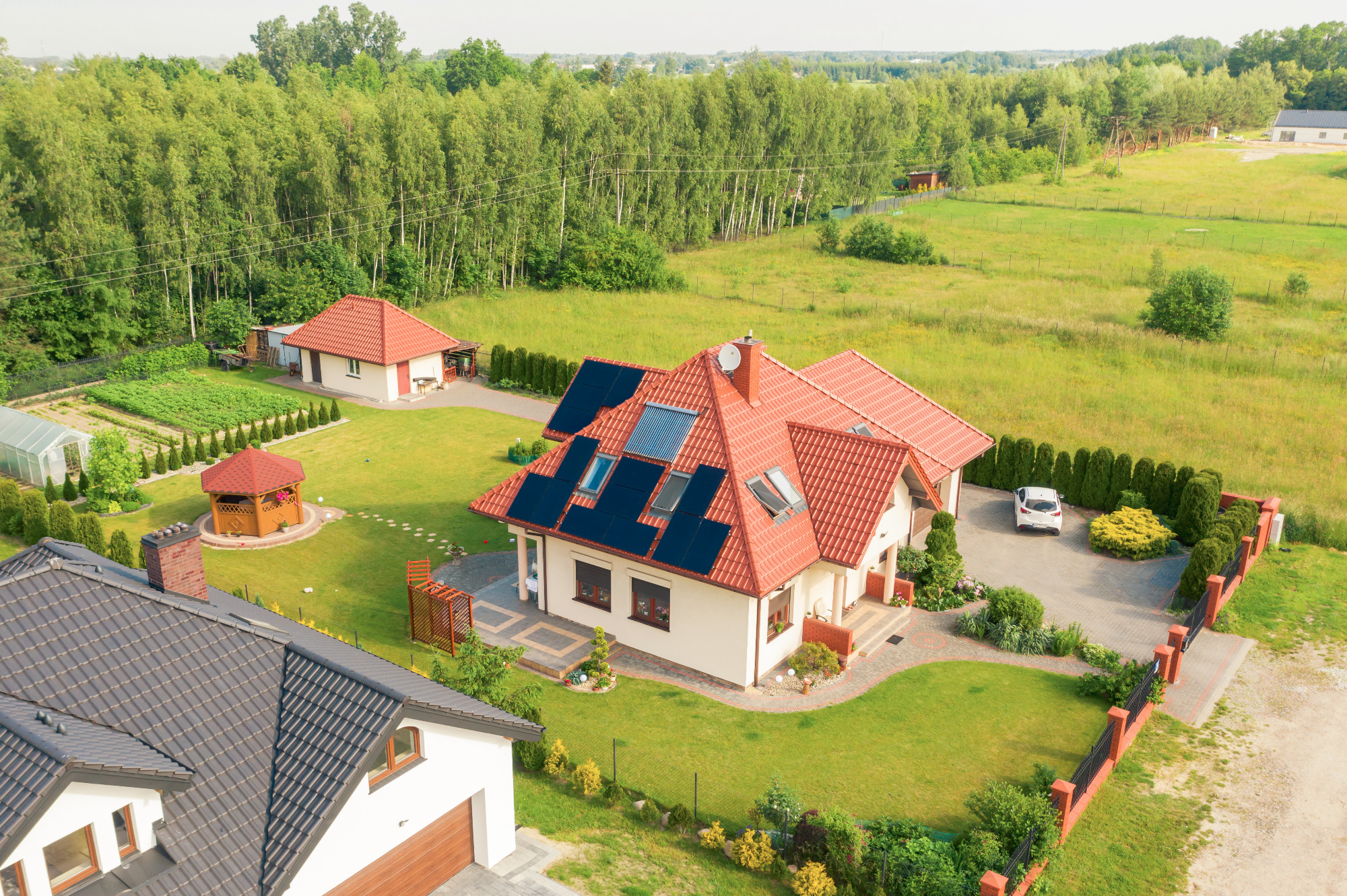 Widok z lotu ptaka na dom z panelami słonecznymi na dachu, czerwona dachówka, zadbany ogród, biały samochód na podjeździe, w tle las.