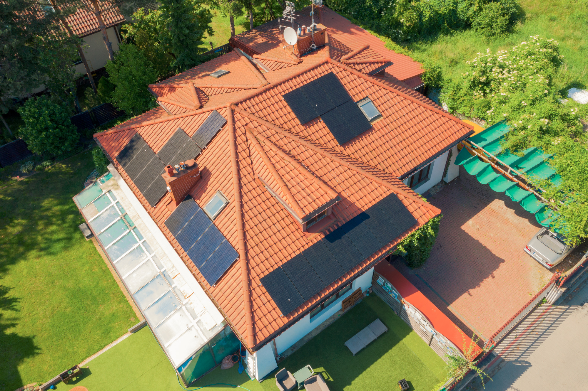 Widok z góry na dom z czerwoną dachówką, na której zamontowane są czarne panele słoneczne; obok domu znajduje się zadaszony taras i zaparkowany samochód.