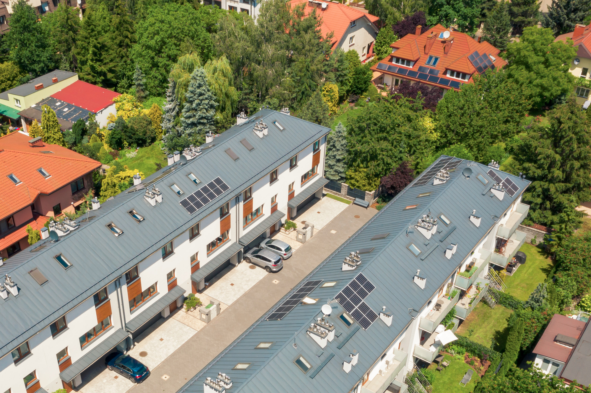 Widok z lotu ptaka na szereg domów z panelami słonecznymi na dachach, otoczonych zielenią i innymi budynkami.