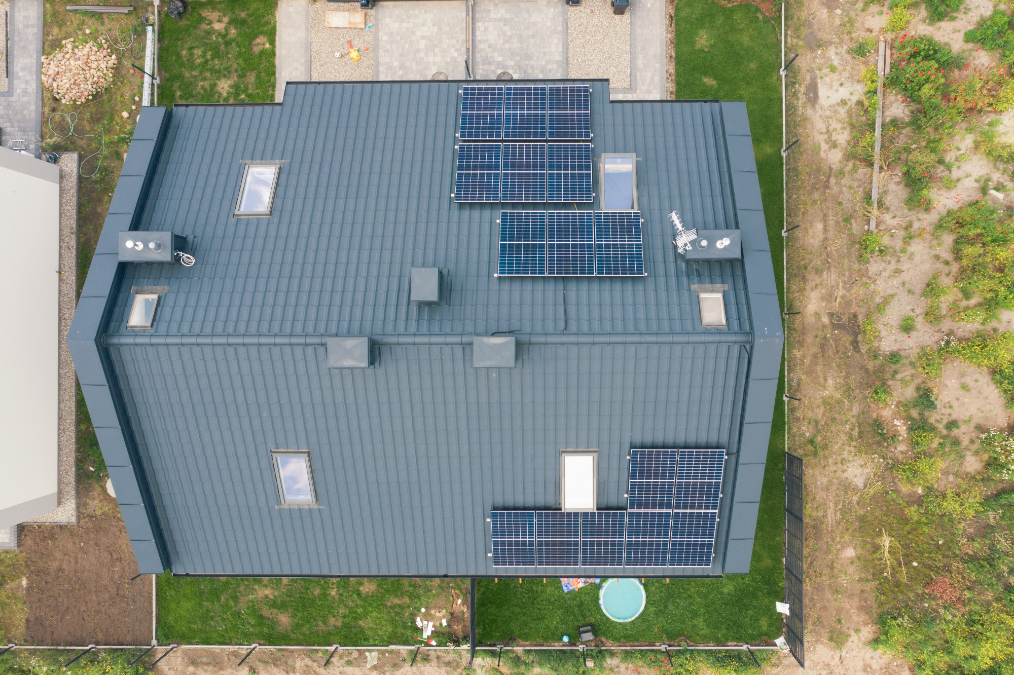 Widok z góry na dach domu z szarej blachodachówki z zamontowanymi panelami fotowoltaicznymi, oknami dachowymi i kominkami wentylacyjnymi.