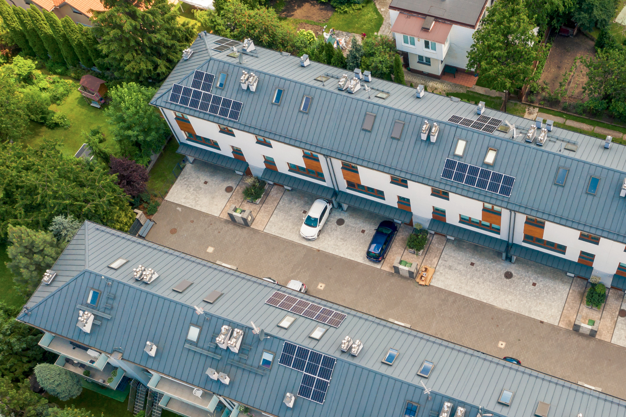 Widok z drona na budynek wielorodzinny z szarym dachem i panelami słonecznymi; na podwórku zaparkowane dwa samochody.