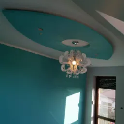 Turkusowa ściana i fragment sufitu z dekoracyjnym podwieszanym elementem w kształcie łzy, oświetlonym nowoczesnym żyrandolem z kryształkami. Podłoga zabezpieczona folią malarską.