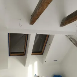 Białe ściany i sufit na poddaszu z drewnianymi belkami, okna dachowe zabezpieczone niebieską taśmą malarską, widoczne przygotowania do malowania.