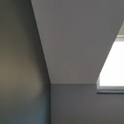 Pomieszczenie po malowaniu: ściana w odcieniach szarości, biały sufit skośny z oknem w tle, zabezpieczona podłoga.
