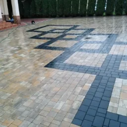 Ułożona kostka brukowa w geometryczny wzór z ciemnoszarymi i beżowymi elementami, mokra powierzchnia odbija światło, w tle widoczny budynek i rząd tuj.