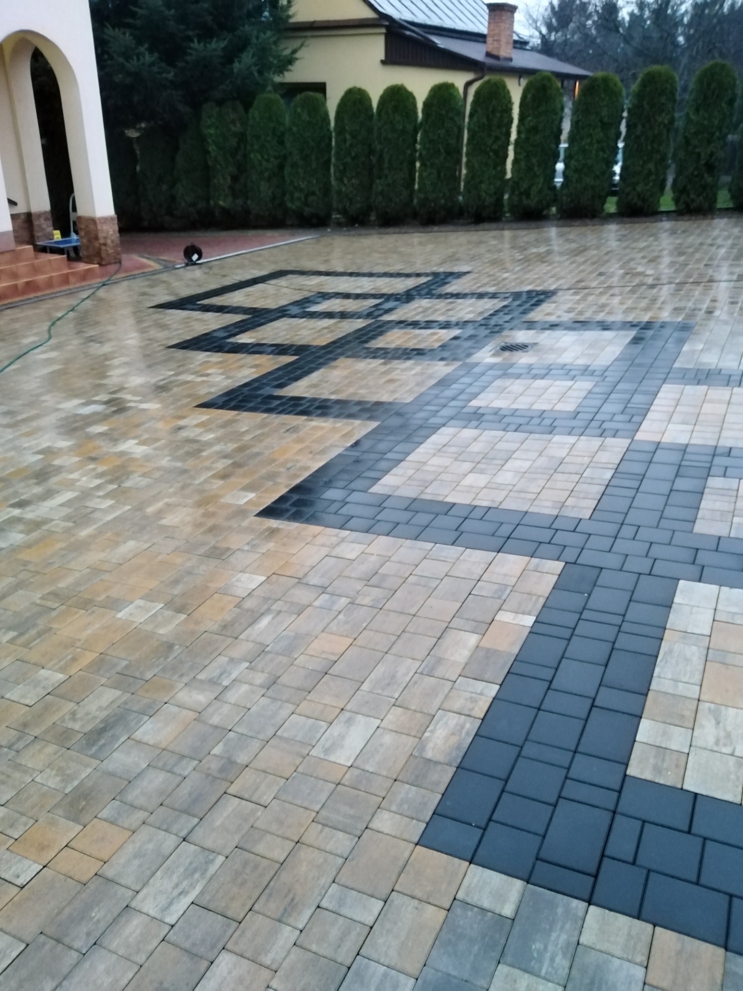 Ułożona kostka brukowa w geometryczny wzór z ciemnoszarymi i beżowymi elementami, mokra powierzchnia odbija światło, w tle widoczny budynek i rząd tuj.
