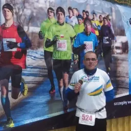 Mężczyzna z medalem na tle baneru z biegnącymi biegaczami, w tle hala sportowa.