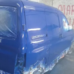 Świeżo polakierowany na niebiesko samochód dostawczy, zabezpieczony folią i papierem malarskim, widoczny na tle ściany z napisem 'MARCEL AUTO'.