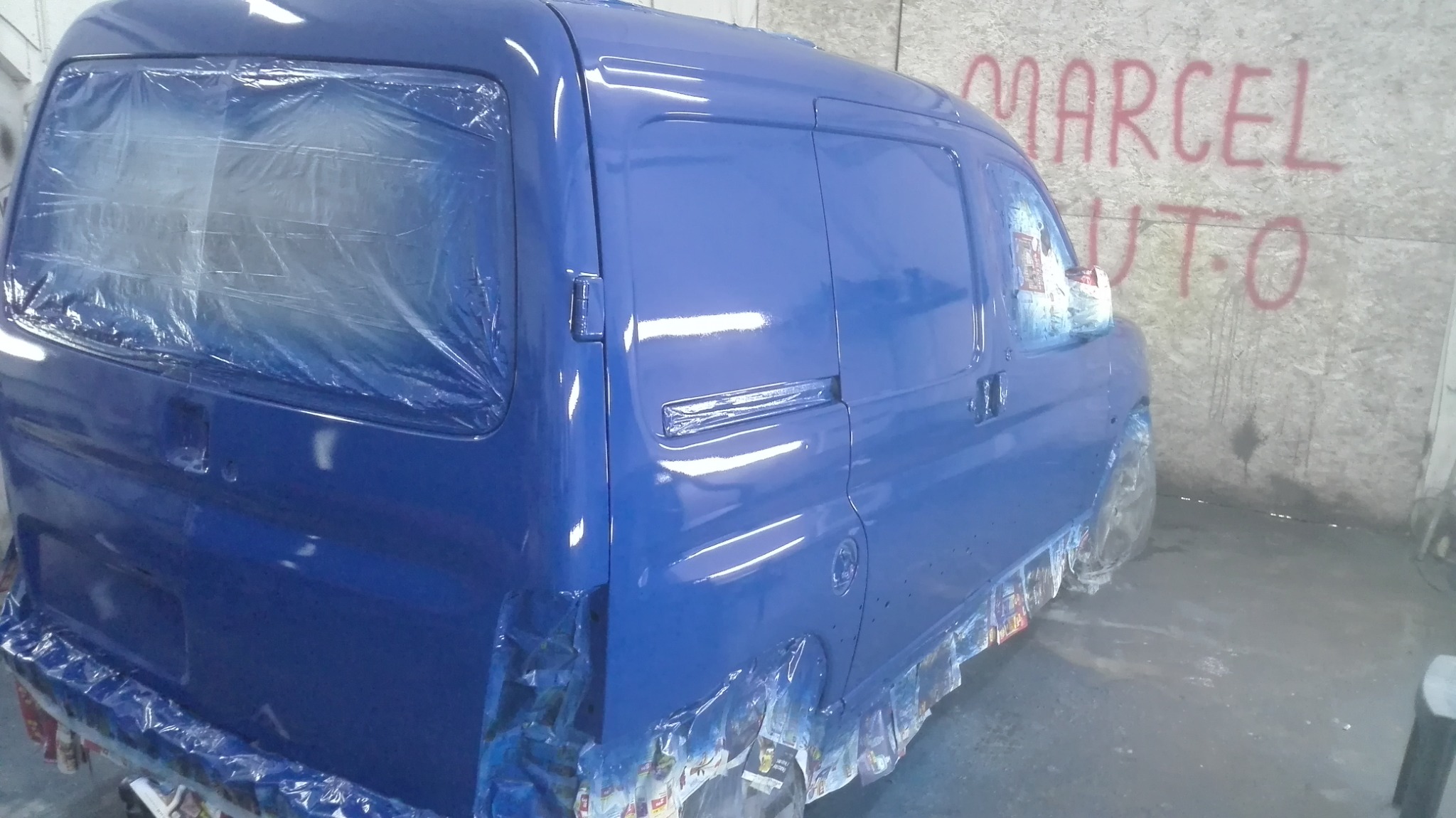 Świeżo polakierowany na niebiesko samochód dostawczy, zabezpieczony folią i papierem malarskim, widoczny na tle ściany z napisem 'MARCEL AUTO'.