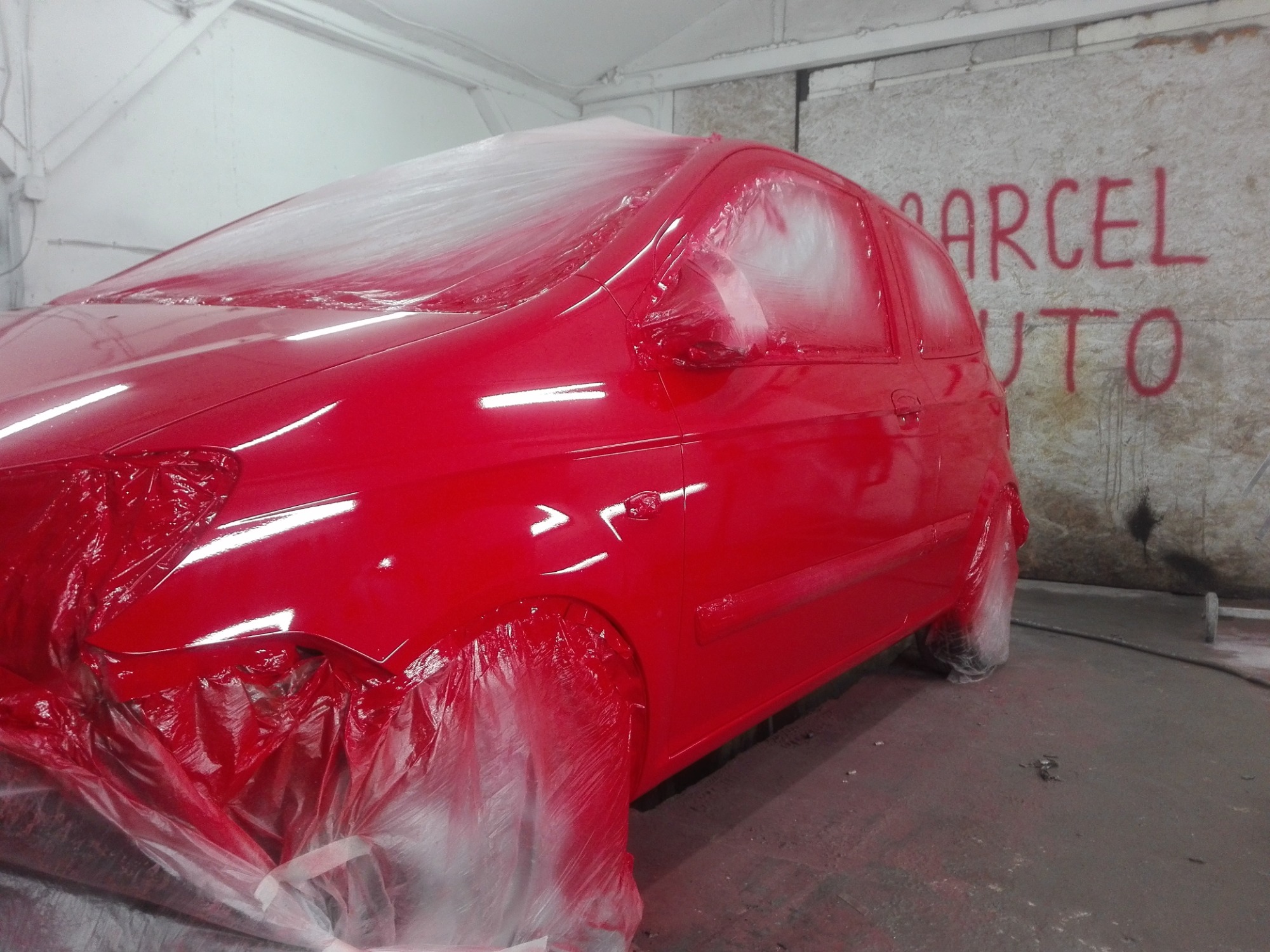 Czerwony samochód osobowy częściowo zakryty folią ochronną w trakcie lakierowania w warsztacie, ściana z napisem w tle.