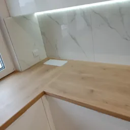 Nowoczesna kuchnia z drewnianym blatem, białymi szafkami i marmurowymi płytkami na ścianie, oświetlona listwą LED pod szafkami górnymi.