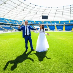 Para młoda w strojach ślubnych pozuje na zielonej murawie stadionu Śląskiego w Chorzowie, w tle widoczne niebieskie trybuny pod białym zadaszeniem.