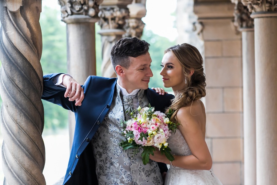 Para młoda w plenerze ślubnym w Mosznej: panna młoda w białej sukni trzyma bukiet kwiatów, pan młody w granatowym fraku opiera się o kolumnę zamku, oboje patrzą na siebie z uśmiechem.