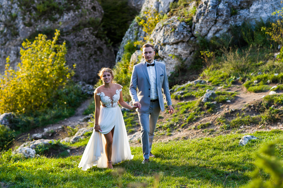 Para młoda idzie po trawiastym wzgórzu w kierunku aparatu, trzymając się za ręce; panna młoda ma białą suknię z koronkową górą i rozcięciem na nodze, a pan młody ma szary garnitur z muchą, w tle...